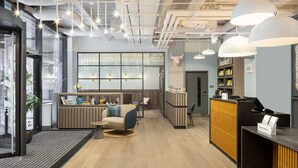 Un espace d’accueil moderne, doté d’un comptoir de réception en bois, d’un coin salon avec un canapé et des fauteuils, et d’une grande fenêtre offrant une vue sur la ville.