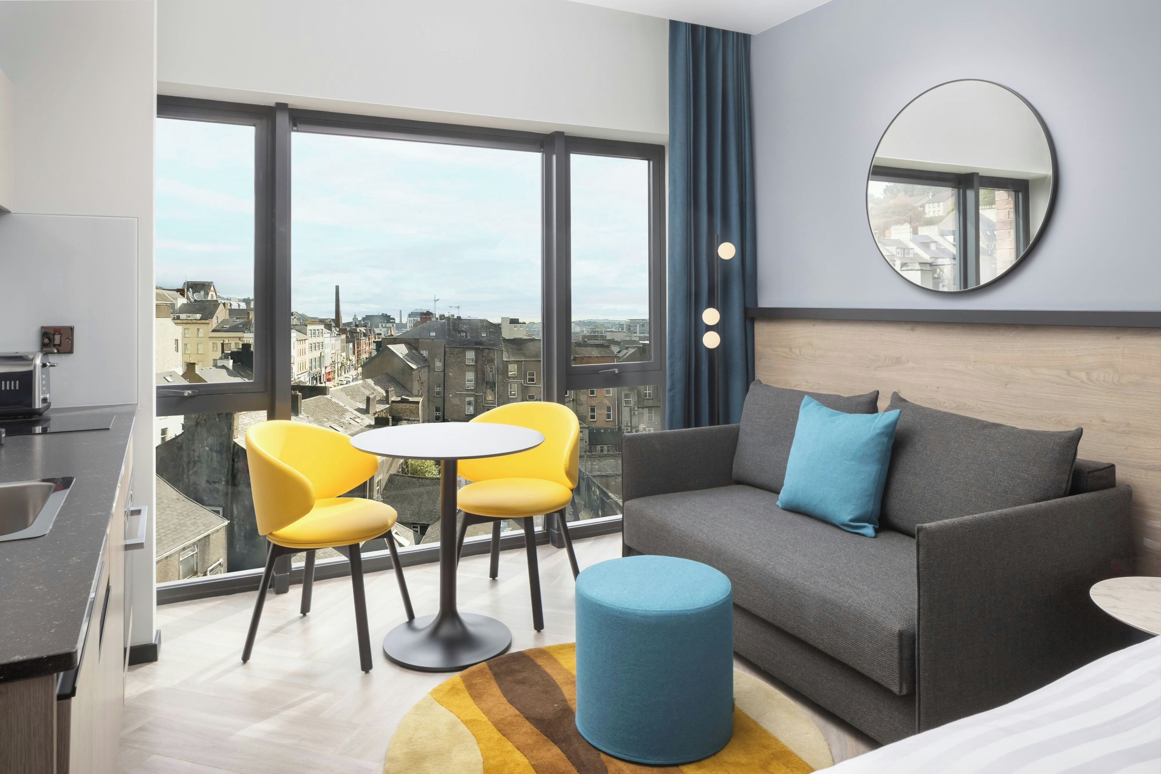 Quarto de hotel moderno com varanda, sofá, duas cadeiras amarelas, mesa redonda, banquinho azul e um espelho grande.