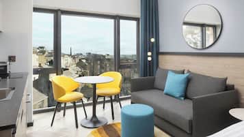 Una habitación de hotel moderna con balcón, un sofá, dos sillones amarillos, una mesa redonda, un reposapiés azul y un espejo grande.