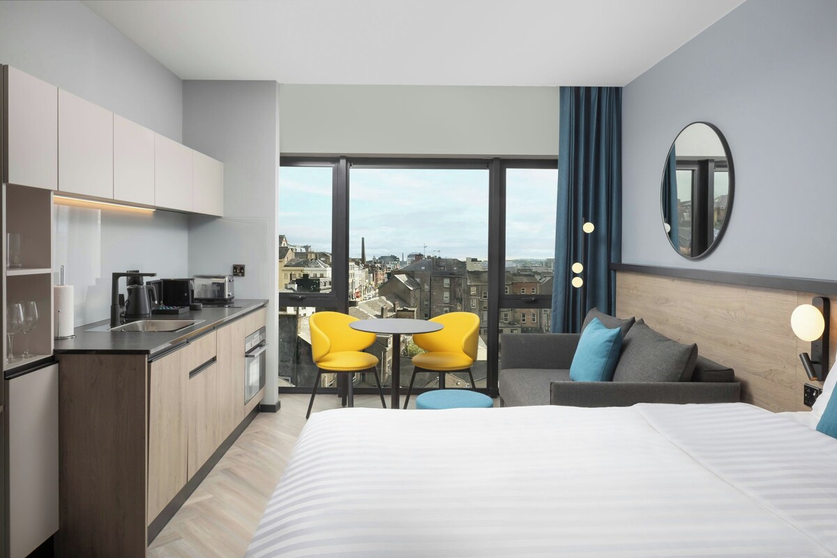 A modern hotel room with a kitchenette, a bed, a seating area with yellow chairs, and a large window offering a city view.