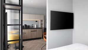 Une chambre d’hôtel moderne équipée d’un lit, d’un téléviseur fixé au mur, d’un coin cuisine avec des placards en bois et d’un coin repas avec une chaise jaune.