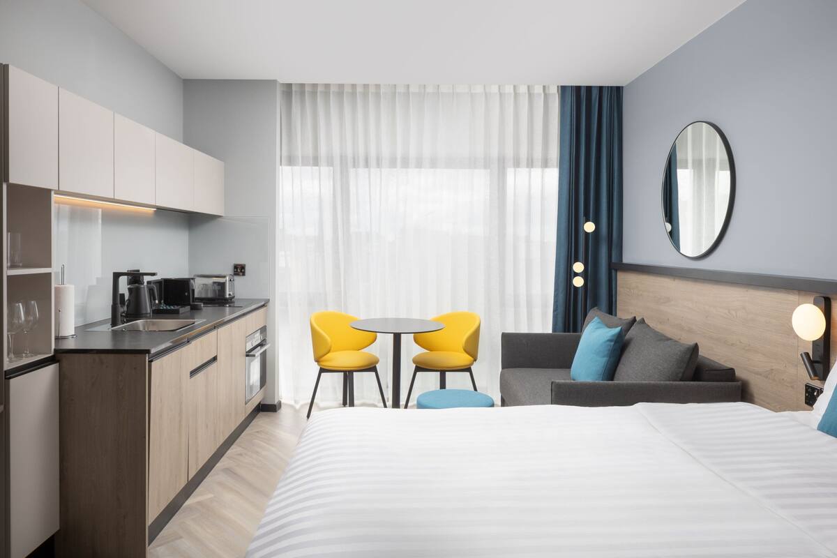 Ein modernes Hotelzimmer mit Bett, einer Couch, einem kleinen Tisch mit Stühlen, einer Küchenzeile und einem großen Fenster mit Vorhängen.