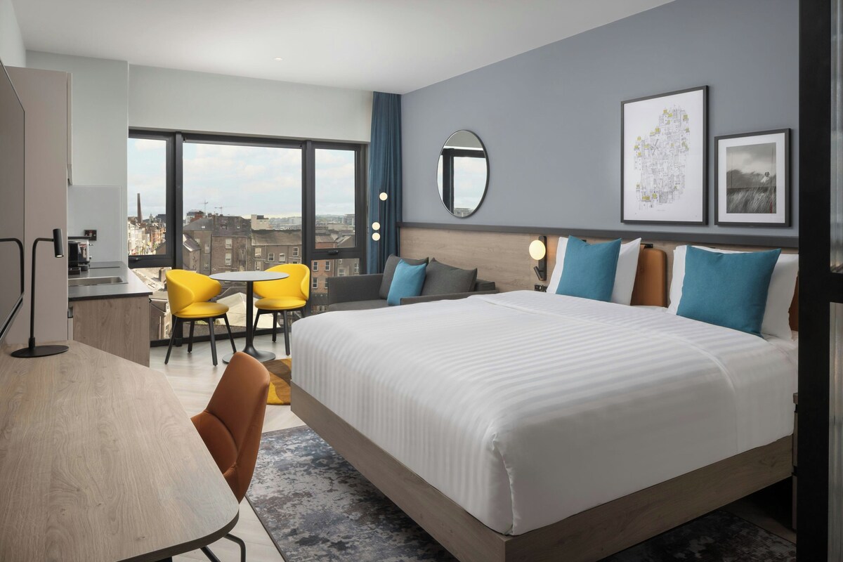 A modern hotel room with a large bed, a desk, and a chair. There is a view of the city through large windows.