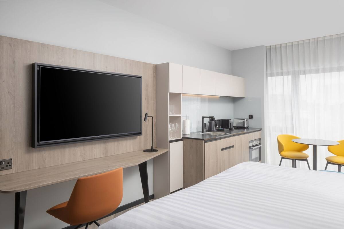 Ein modernes Hotelzimmer mit Flachbildfernseher, einem Holztisch, einem Bett, einer kleinen Küchenzeile und einem Essbereich mit gelben Stühlen.