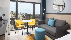 Une chambre moderne avec un balcon, un canapé gris, des fauteuils jaunes, un miroir rond et une vue sur la ville.