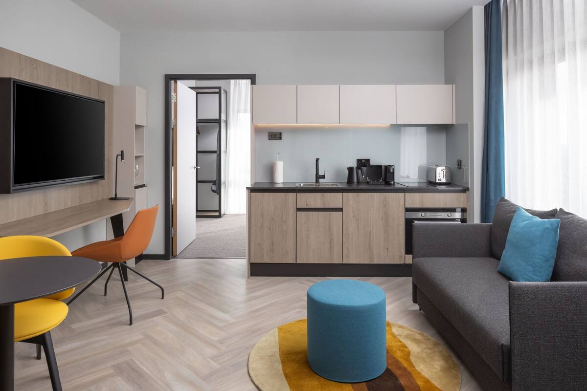 Ein modernes Wohnzimmer mit Sofa, Hocker und Essbereich mit Stühlen. Im Hintergrund ist eine Küche mit Schränken und Geräten zu sehen.