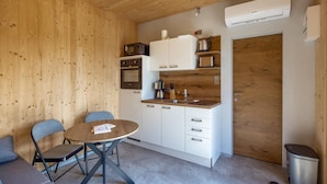 Una cocina compacta con armarios blancos, un microondas, una cafetera y un lavamanos. Hay una mesa redonda con dos sillas y un cubo de basura.