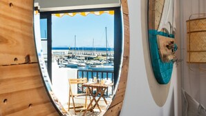 On aperçoit, à travers une fenêtre à cadre en bois, un port de plaisance avec des bateaux, une table entourée de chaises et une planche de surf accrochée au mur.