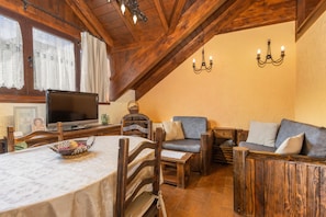 Ein gemütliches Zimmer mit Esstisch, Holzstühlen, einer Couch, einem Fernseher und einem Chandelier.