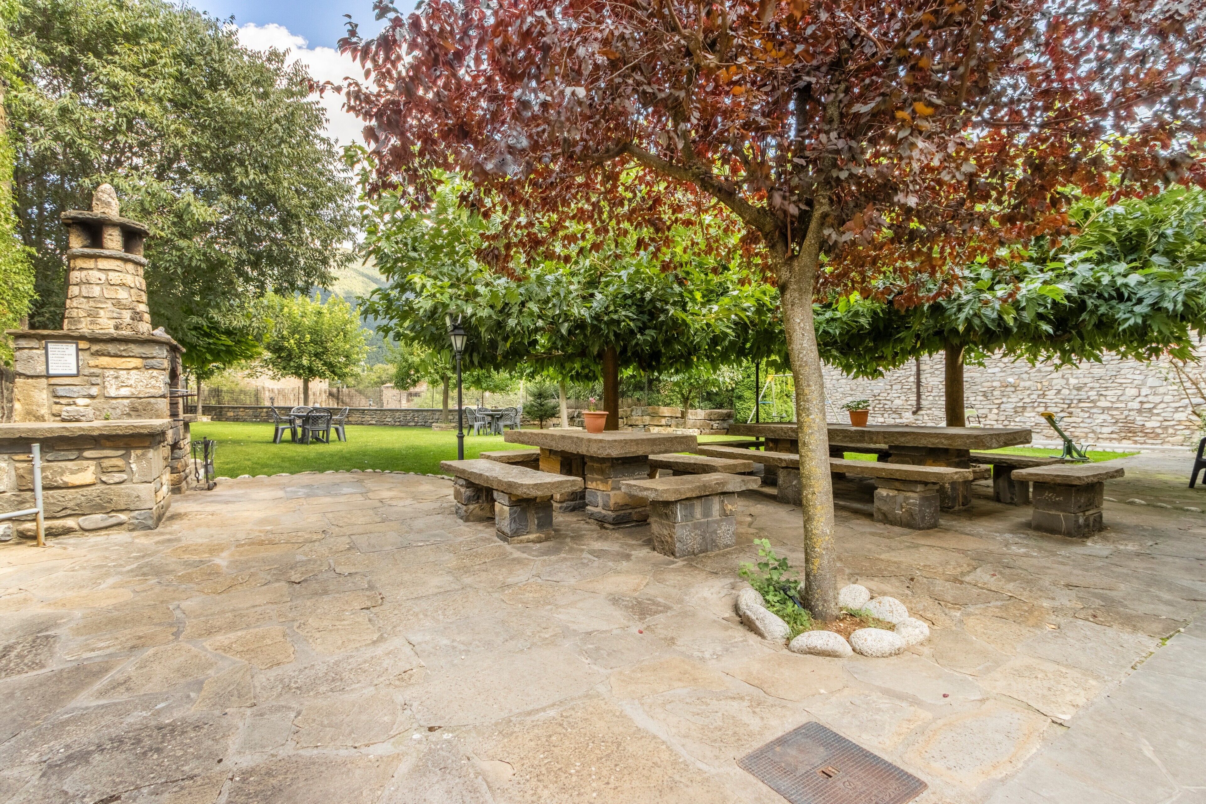 Una veranda esterna con tavoli e panche in pietra, circondata da alberi e un muro di pietra.