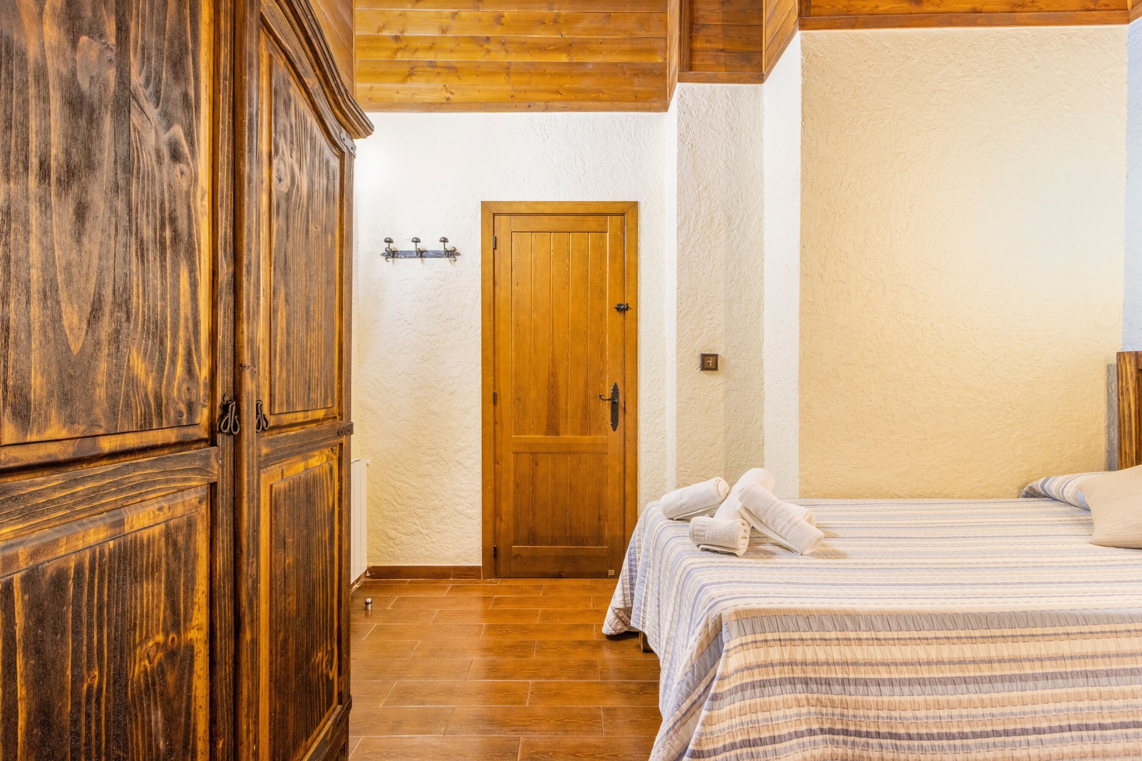 Quarto com guarda-roupa de madeira, uma porta, cama com colcha listrada e parede branca.