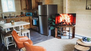 A cozy kitchen with wooden cabinets, a stainless steel refrigerator, a microwave, and a TV displaying a fire. There is a dining table with chairs and a small round table with a plant. - Raven's Spruce Suite #7 Getaway (Fairbanks)