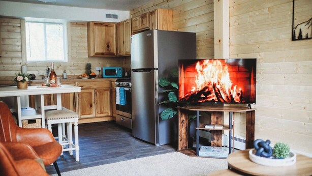 A cozy cabin-style kitchen with wooden cabinets, a stainless steel refrigerator, a microwave, and a large TV displaying a fireplace. - Raven's Spruce Suite #7 Getaway (Fairbanks)