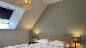 Une chambre avec un plafond en pente, un puits de lumière, un lit avec une literie blanche, deux tables de chevet avec des lampes et une tête de lit en bois.