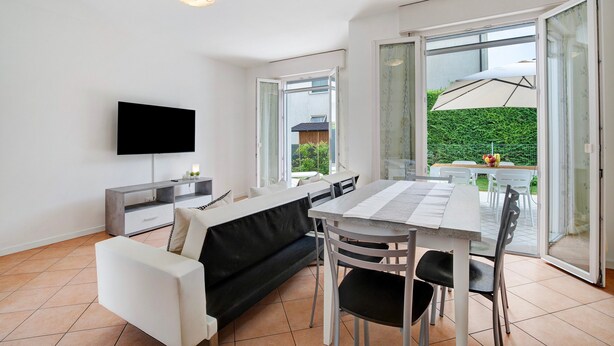 Una sala moderna con un televisor de pantalla plana, un sofá, una mesa de comedor con sillas, y un ventanal corredizo que da a un área exterior con mesa y sillas.