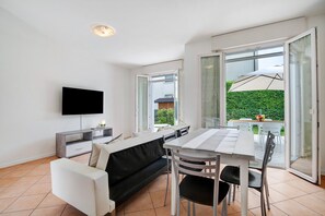 Una sala moderna con un televisor de pantalla plana, un sofá, una mesa de comedor con sillas, y un ventanal corredizo que da a un área exterior con mesa y sillas.