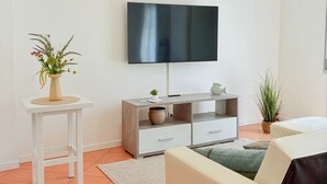 Una sala moderna con un televisor de pantalla plana, un sofá blanco, una mesita auxiliar con un florero y un mueble gris con cajones.