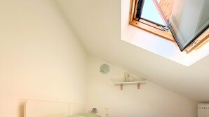Un dormitorio con cama, mesita de noche, lámpara, estante con objetos decorativos y tragaluz.