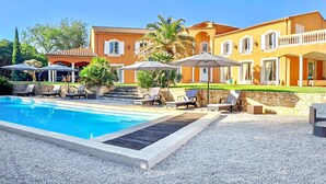 A swimming pool with lounge chairs and umbrellas, surrounded by a stone wall and a building with a tiled roof.