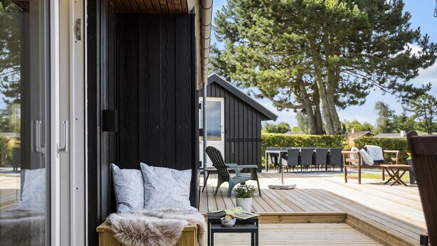 A wooden deck with a seating area, a small table with a bowl, and a view of a building and trees. - Awesome home in Bogense with WiFi (Bogense)