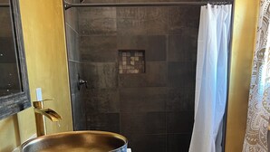Un baño con un recipiente decorativo grande sobre el lavamanos, una ducha con cortina blanca y suelo de madera.