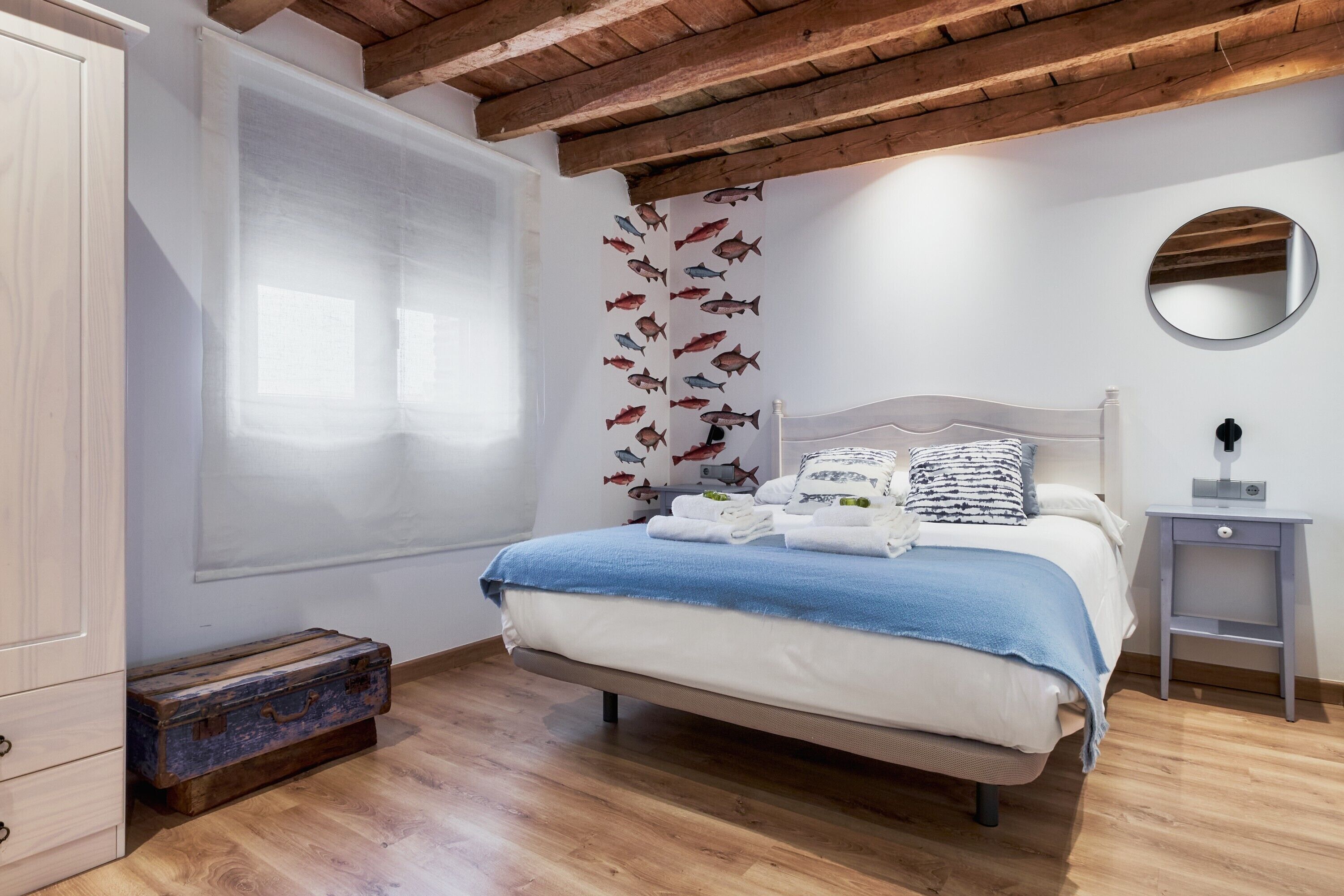 Un dormitorio con cama, techo de madera, ventana con persianas, pared decorada con calcomanías de peces, mesita de noche y maleta.