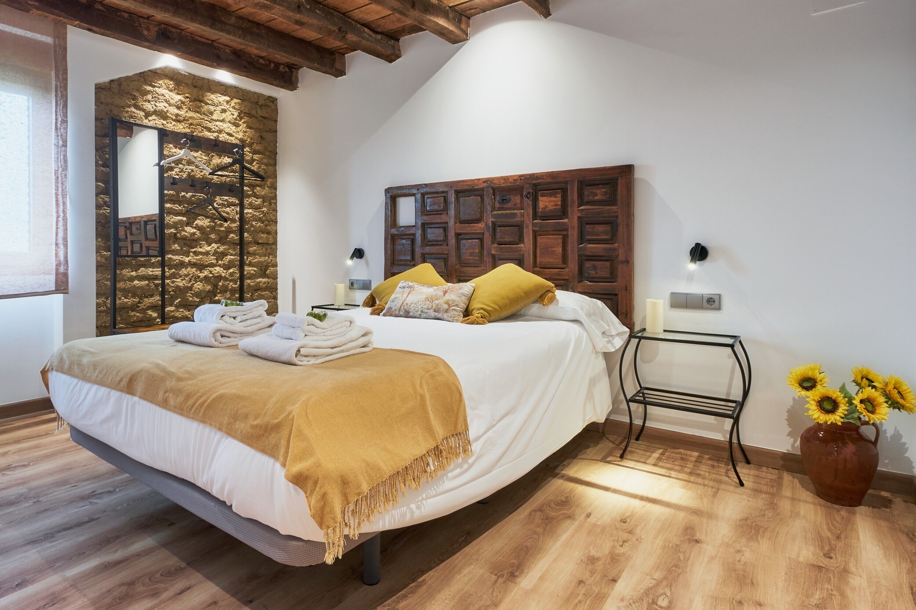 Un dormitori amb un llit, un capçal de fusta, una taula de nit, una prestatgeria penjada a la paret i un vas amb gessamins.