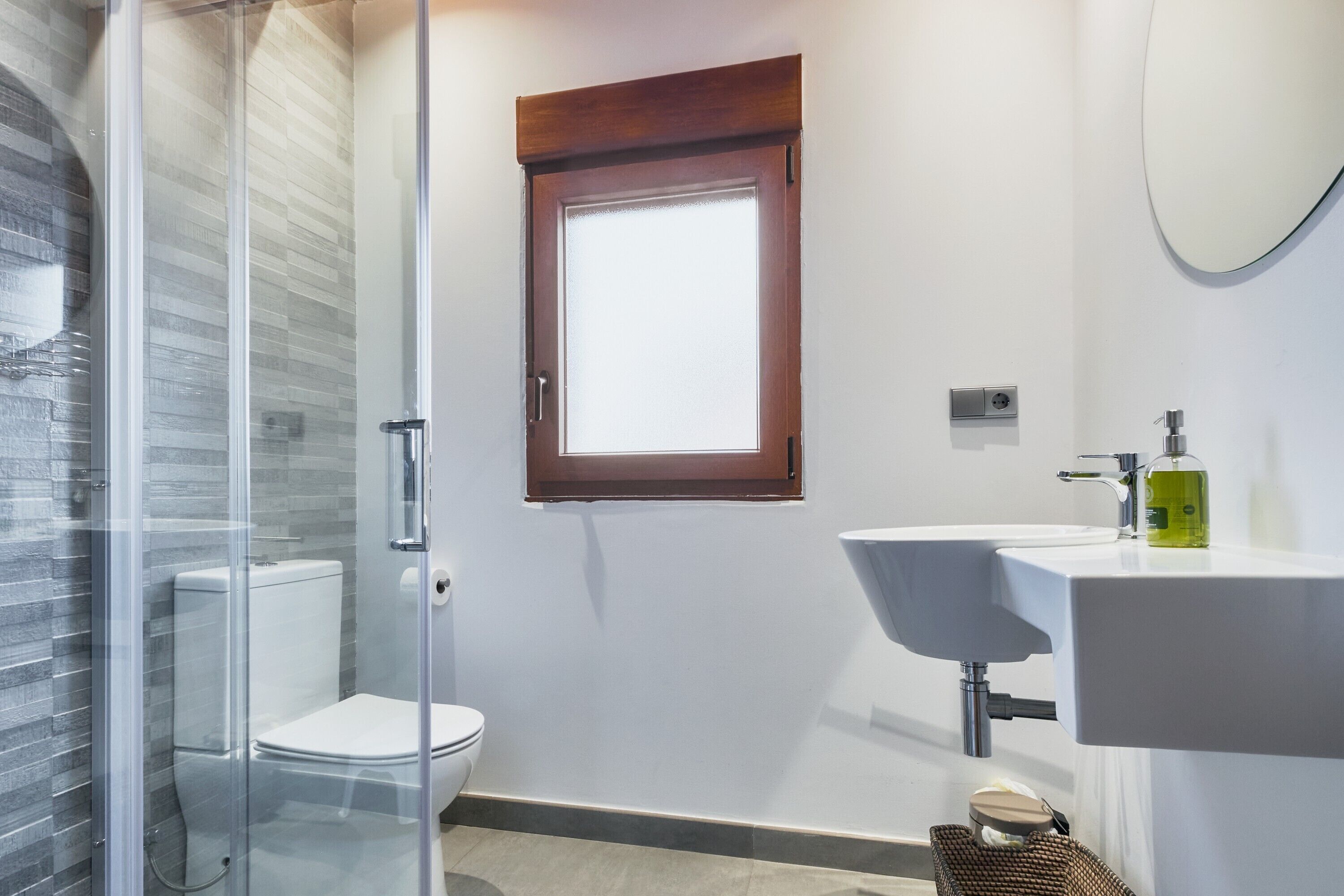 Un baño moderno con cabina de ducha de vidrio, inodoro, lavabo con espejo y ventana sobre el lavabo.