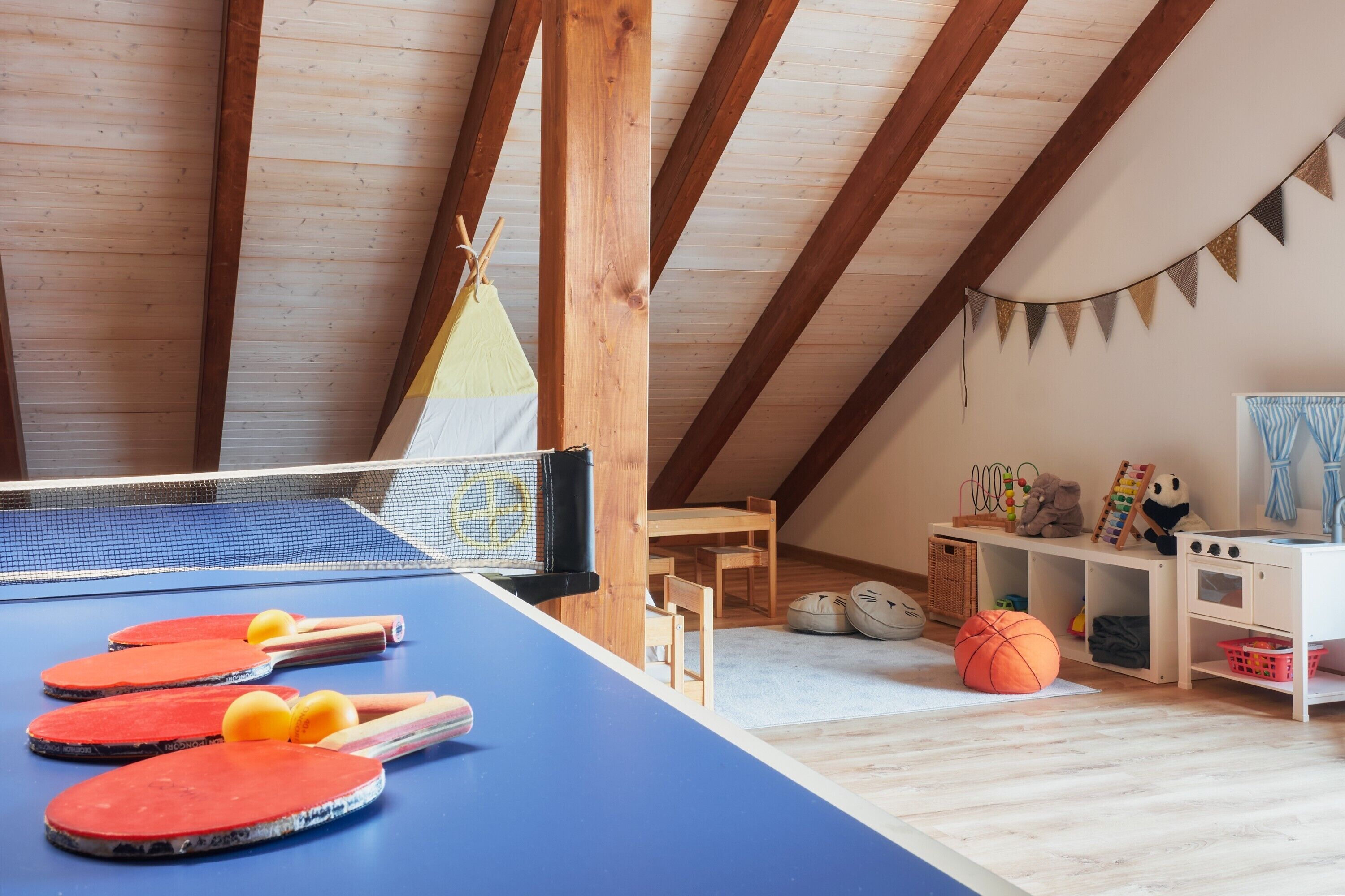 Ein Zimmer mit Tischtennisplatte, Holzmöbeln, einer kleinen Spielecke und einer erhöhten Decke.