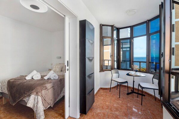 Een slaapkamer met een bed, een kleine tafel met stoelen, een raam met uitzicht op zee en een deur die naar een andere kamer leidt.