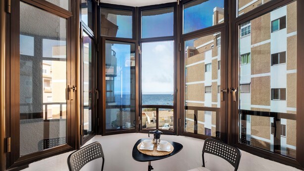 A corner balcony with two chairs, a small table, and a view of the sea and buildings.