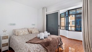 A bedroom with a bed, a nightstand, a window with curtains, and a view of buildings outside.