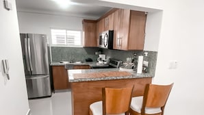 A modern kitchen with a stainless steel refrigerator, wooden cabinets, and a granite countertop. There are two wooden bar stools with cushioned seats. - Senderos del botánico  (Santiago de los Caballeros)