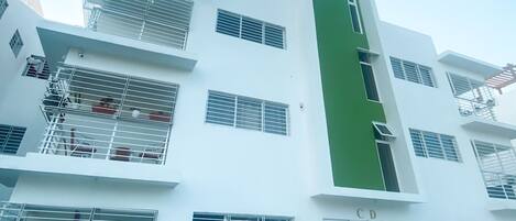 A multi-story building with white exterior walls, multiple balconies with metal railings, and a green vertical stripe on one side.
