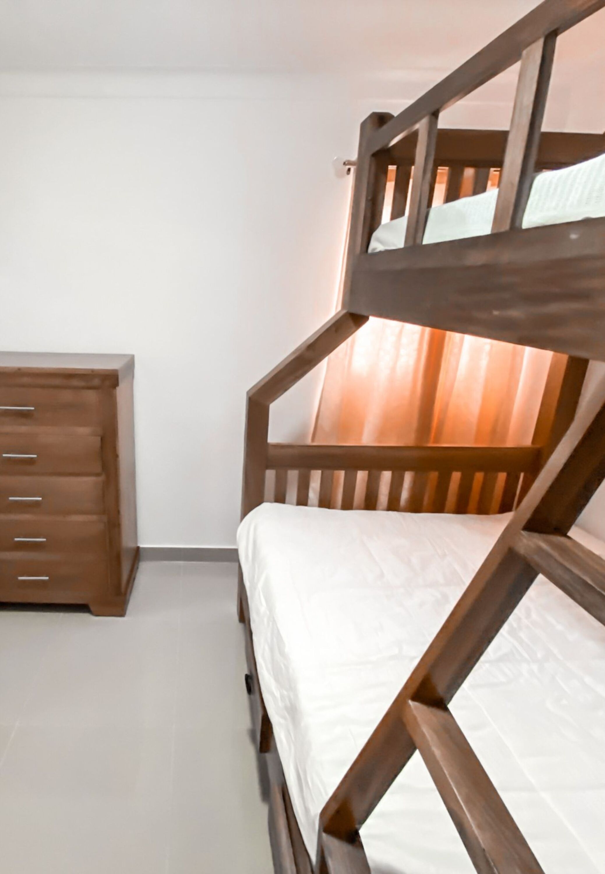 Ein Etagenbett mit Holzrahmen und weißer Bettwäsche, eine Holzkommode mit Schubladen und ein fensterbedeckender Vorhang.