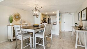 Un coin repas avec une table en bois et des chaises blanches, un lustre et un miroir en forme de soleil accroché au mur.