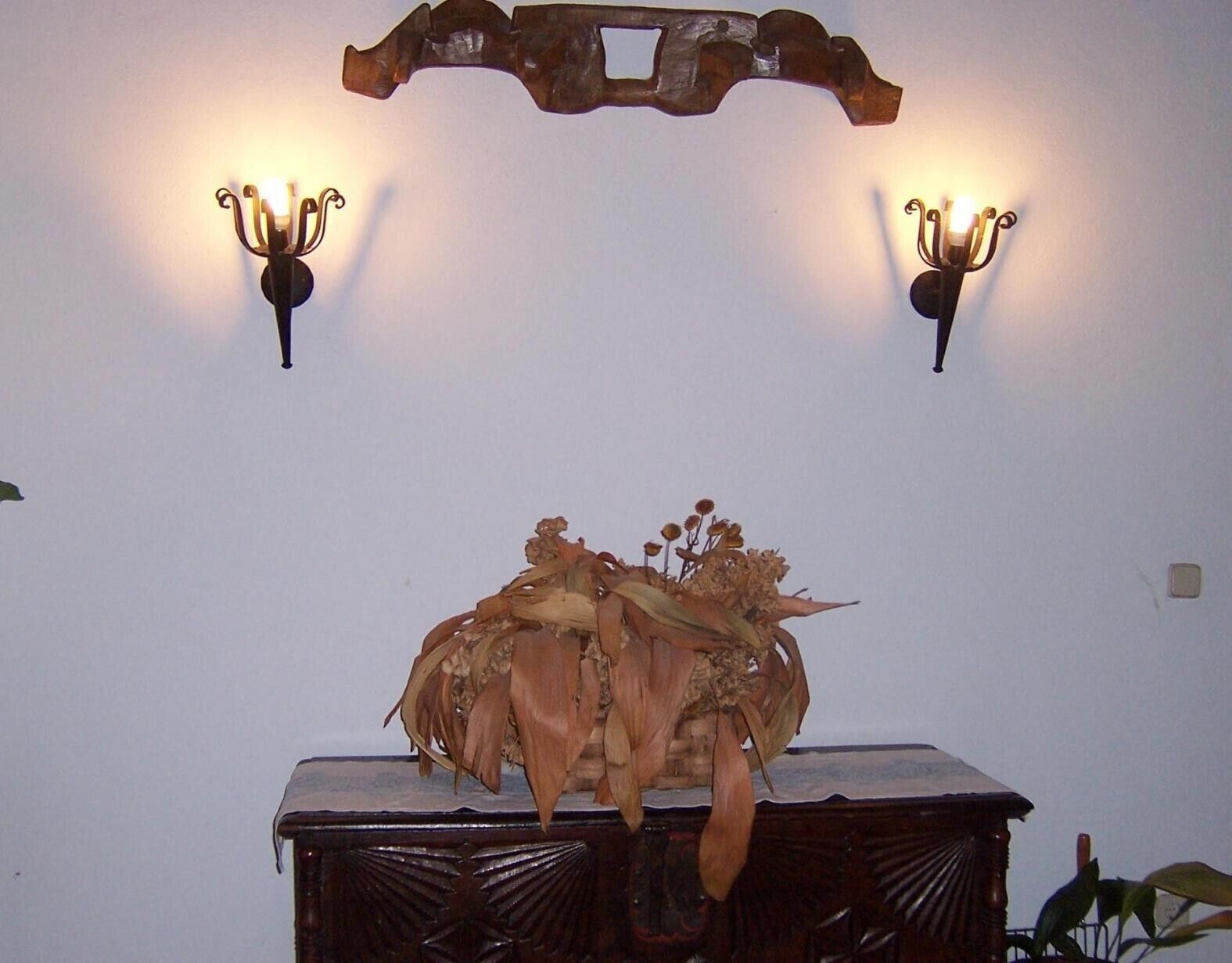 Una habitación con una escultura de madera decorativa sobre una mesa, dos lámparas fijadas a la pared y un elemento de madera en la pared encima de la escultura.