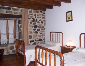 Quarto com parede de pedra, estrutura de cama de madeira, mesa de cabeceira pequena com abajur e quadro pendurado na parede.