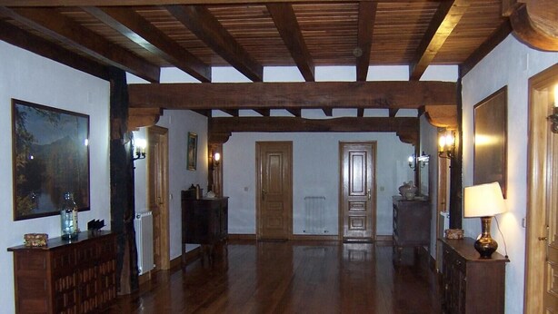 Een ruime kamer met houten vloeren, een hoge plafond met zichtbare balken, een groot schilderij aan de linkerkant en een deur aan het einde.