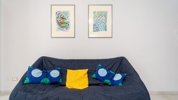 Un canapé de couleur sombre agrémenté de coussins bleus et jaunes, deux tableaux abstraits encadrés accrochés au mur et un sol carrelé.