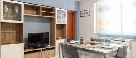 A compact living space with a dining area, built-in shelving, a television, and a window with blinds.