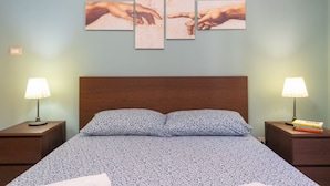 Un lit avec une couvre-lit à motifs, deux lampes de chevet, une tête de lit en bois et une œuvre d’art représentant des mains, accrochée au mur.