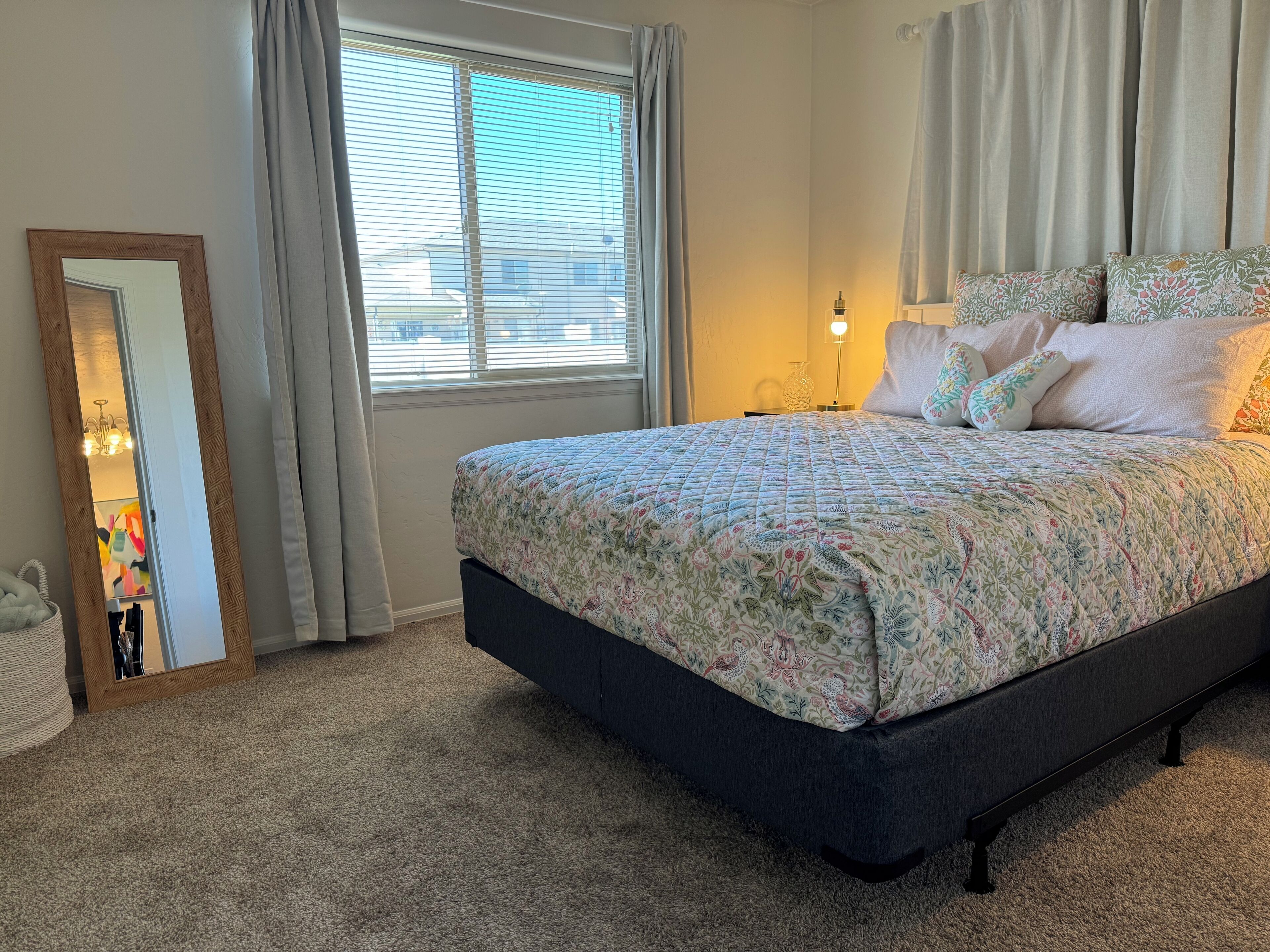 A bedroom with a large bed, a mirror, a window with curtains, and a nightstand with a lamp.