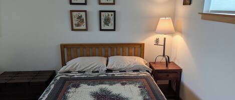 A bedroom with a wooden bed, a nightstand with a lamp, and four framed botanical prints on the wall.