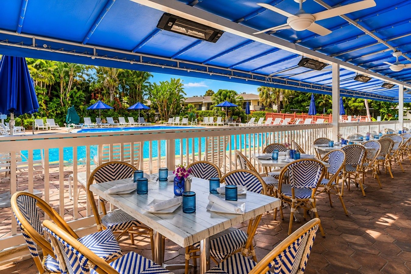 An outdoor pool area with lounge chairs and tables, a pool, and a covered seating area with umbrellas.