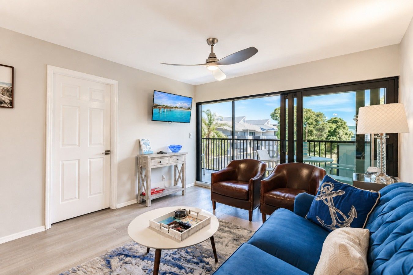 A living room with a ceiling fan, a television, a sofa with decorative pillows, a coffee table with magazines, and a view of the outside through large windows.