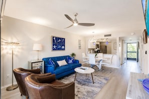 A modern living room with a blue sofa, leather chairs, a dining area, and a kitchen in the background. - Park Shore Resort- BLDG I #351*Remodeled* (Park Shore Resort)