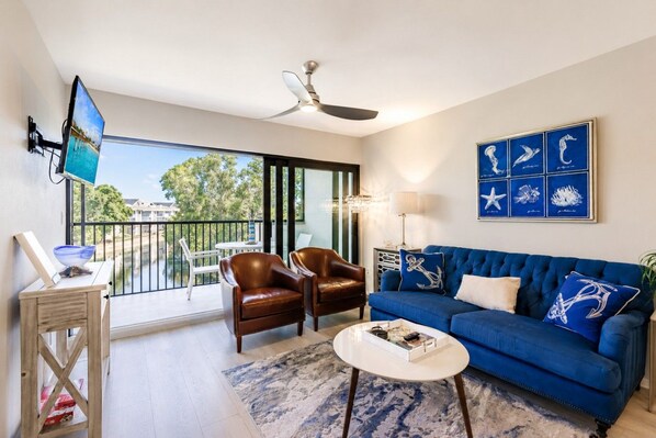 A living room with a blue sofa, two leather chairs, a coffee table, a TV, and a balcony with a view. - Park Shore Resort- BLDG I #351*Remodeled* (Park Shore Resort)