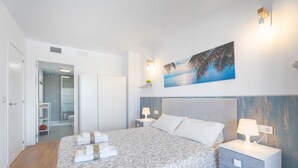 Una habitación de hotel con una cama, una mesita de noche, una lámpara de pared, un perchero y un cuadro de palmeras.