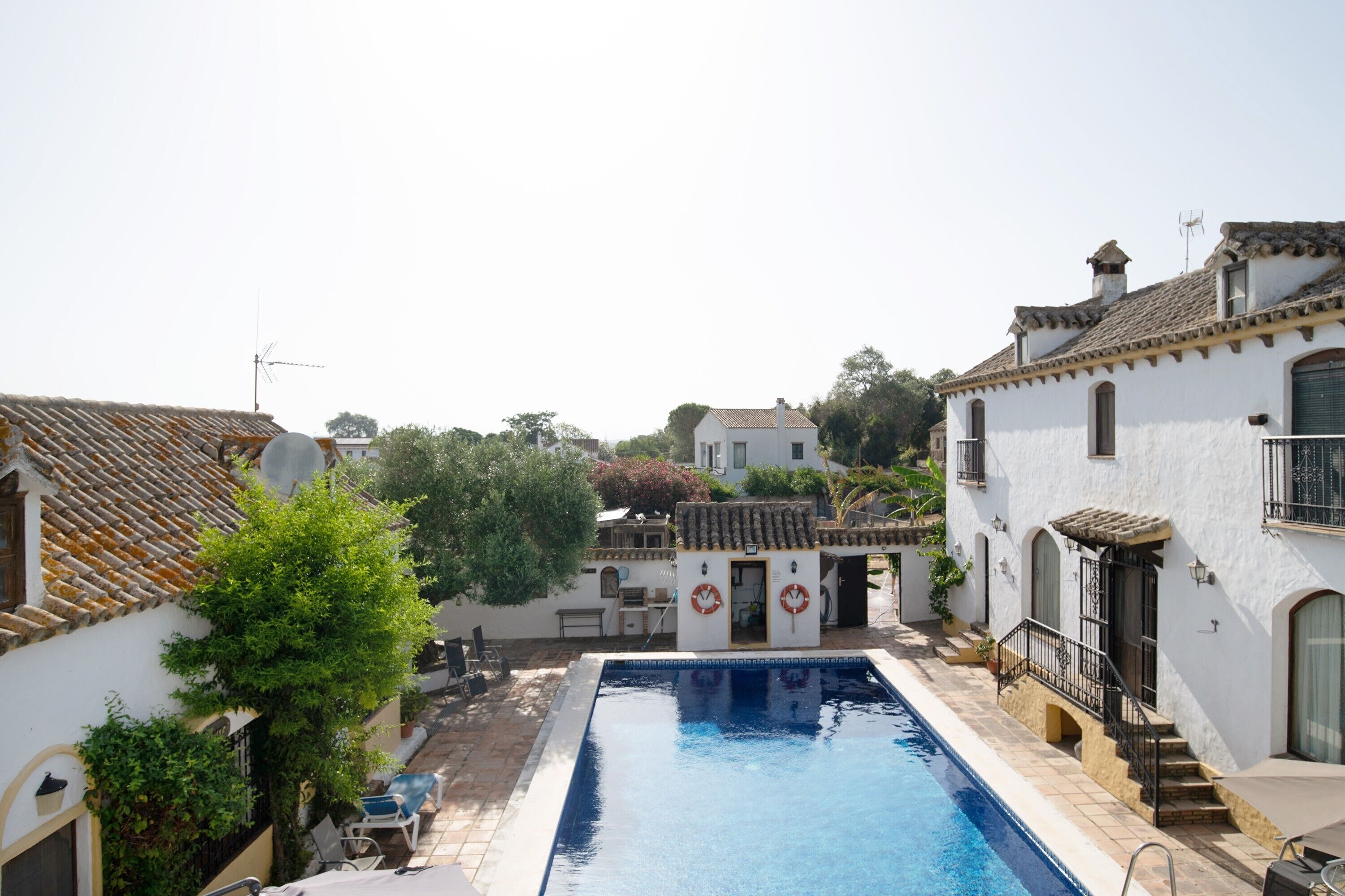 Piscina cercada por edifícios brancos com telhados azulejados, uma pequena estrutura com boia salva-vidas e uma árvore.